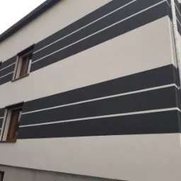 Dwukondygnacyjny dom z kremową elewacją i poziomymi pasami w kolorze antracytowym, malowanymi przez pracownika z użyciem pędzla, widoczne okna z brązowymi ramami i częściowo opuszczonymi roletami.