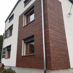 Fragment elewacji domu z brązową okładziną imitującą drewno, widoczne okna z roletami i rynna spustowa, tynk w kolorze złamanej bieli.