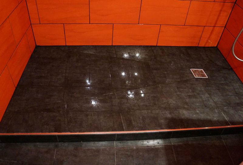 Wykończona kabina prysznicowa z ciemną, błyszczącą posadzką z płytek i pomarańczowymi płytkami ściennymi, widoczny odpływ liniowy i chromowana słuchawka prysznicowa.