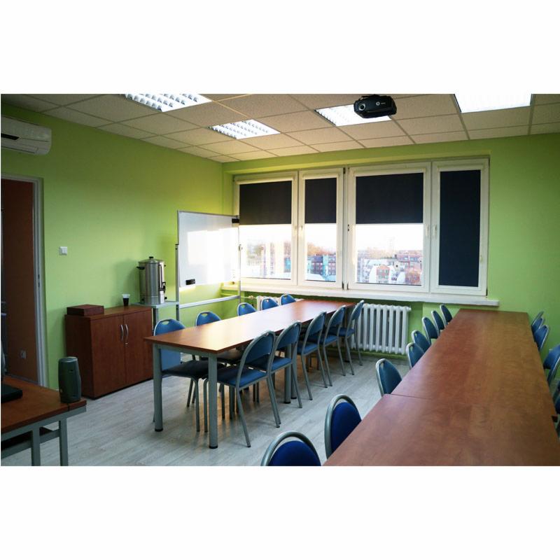 Sala konferencyjna z długim stołem, krzesłami z niebieskim obiciem, zielonymi ścianami i projektorem zamontowanym na suficie podwieszanym z kasetonów.