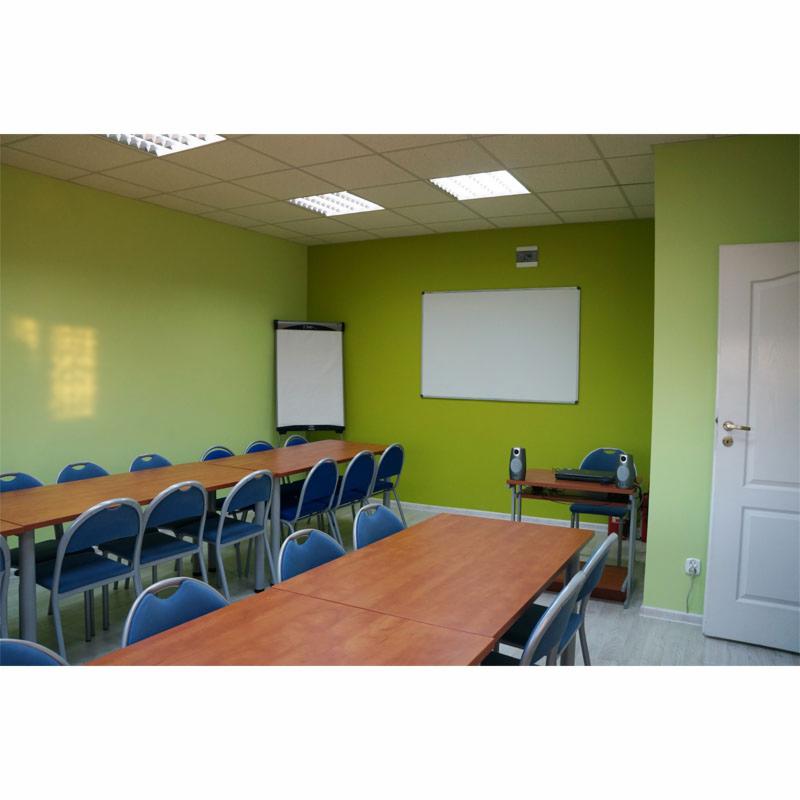 Jasnozielona sala konferencyjna z dwoma długimi drewnianymi stołami, niebieskimi krzesłami, tablicą suchościeralną i flipchartem, oświetlona panelami LED w suficie kasetonowym.
