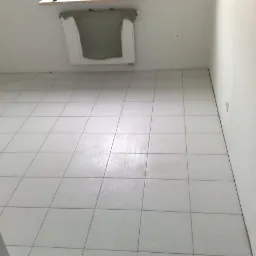 Wyłożona jasnymi, kwadratowymi płytkami podłoga w pustym pomieszczeniu z grzejnikiem, na którym wisi szara koszulka.