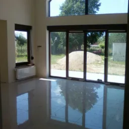 Jasne wnętrze z nową, błyszczącą podłogą z płytek, dużymi oknami z widokiem na ogród i stertę piasku na zewnątrz.