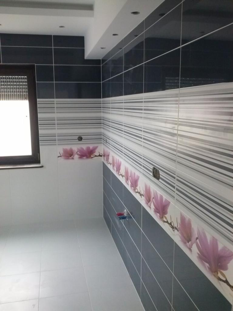 Wykończona łazienka z ciemnymi i jasnymi płytkami, dekoracyjnym pasem z motywem kwiatowym i szarą podłogą, widok z niskiej perspektywy.