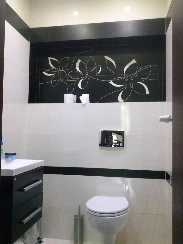 Wykończona łazienka w bieli i czerni z motywem kwiatowym na czarnych płytkach, toaleta, umywalka z szafką i akcesoria łazienkowe.