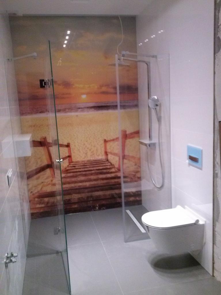 Szklana kabina prysznicowa z motywem plaży i zachodu słońca na ścianie, wisząca toaleta, szare płytki podłogowe i ścienne, nowoczesna aranżacja łazienki.