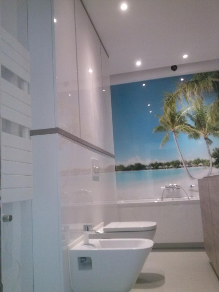 Nowoczesna łazienka z białymi sanitariatami, bidetem i toaletą, na ścianie fototapeta z motywem tropikalnej plaży, oświetlenie punktowe.