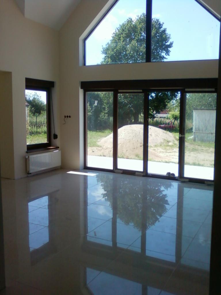 Jasne wnętrze z nową, błyszczącą podłogą z płytek, dużymi oknami z widokiem na ogród i stertę piasku na zewnątrz.