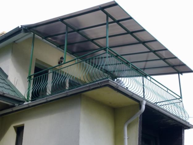Zadaszony balkon z zieloną, metalową balustradą o falistym wzorze, zamontowany na elewacji domu z widocznym fragmentem dachu.