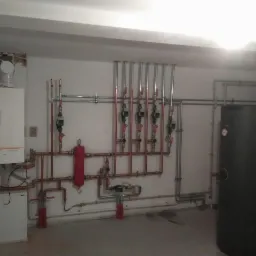 Zainstalowany piec gazowy z rozbudowaną instalacją hydrauliczną, widoczne liczne rury miedziane, zawory i zbiornik akumulacyjny w surowym pomieszczeniu kotłowni z widocznymi śladami po pracach...