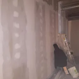 Ściana z płyt kartonowo-gipsowych w trakcie szpachlowania, widoczne łączenia i nierówności, drabina oparta o ścianę, worek gładzi i ubranie robocze w kącie.