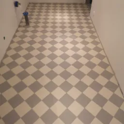 Ukończona podłoga w łazience, wyłożona błyszczącymi, szaro-białymi płytkami w romby, z widocznymi przyłączami hydraulicznymi na ścianie i fragmentem progu drzwiowego.