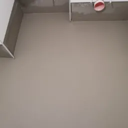 Wyrównana, szpachlowana powierzchnia podłogi w łazience, widoczne rury instalacyjne pod sufitem i fragmenty białych ścian, przygotowane do dalszych prac wykończeniowych.