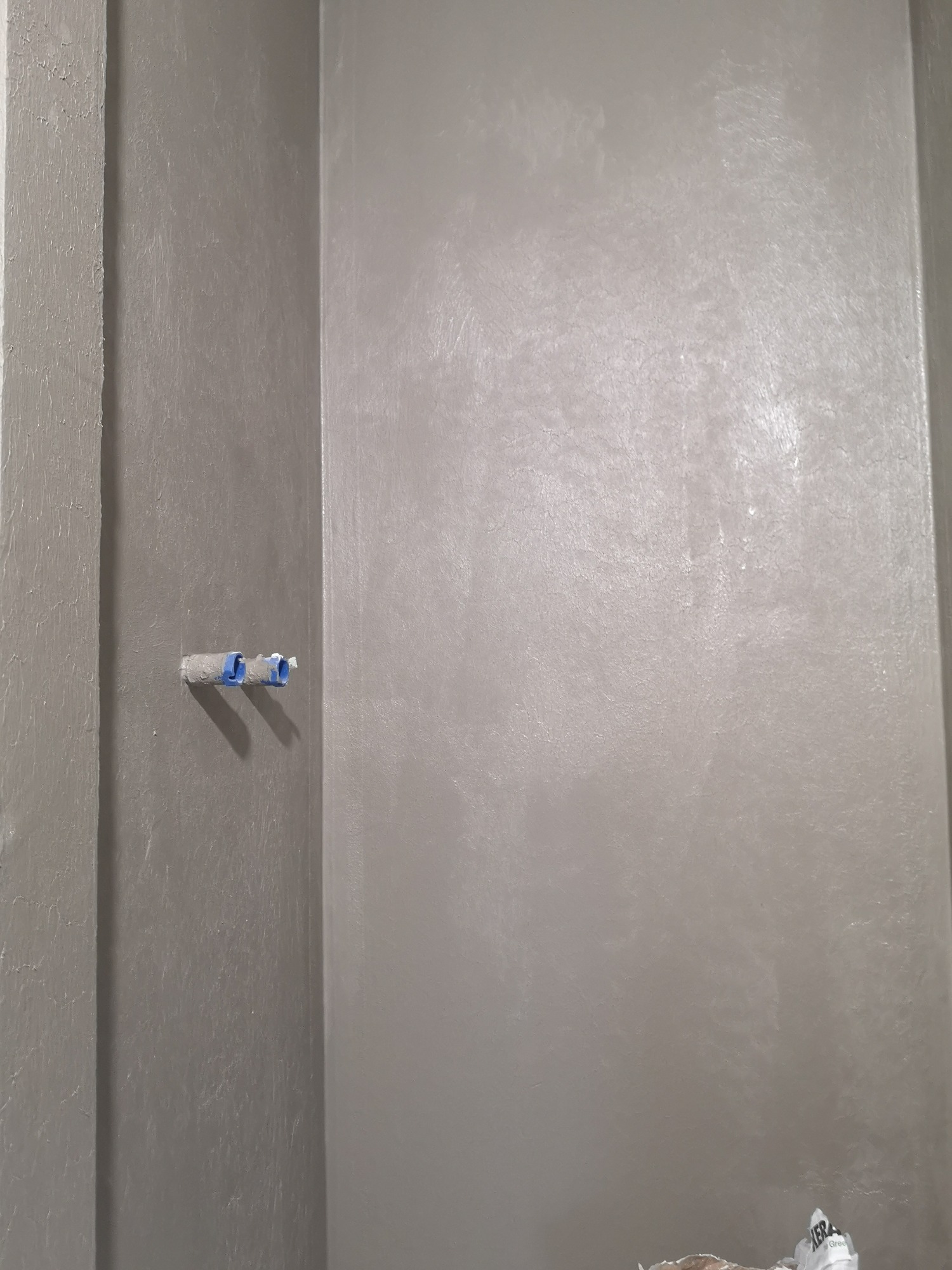 Świeżo wyszpachlowana, szara ściana w łazience z widocznymi rurami instalacji wodnej zabezpieczonymi niebieską taśmą malarską.