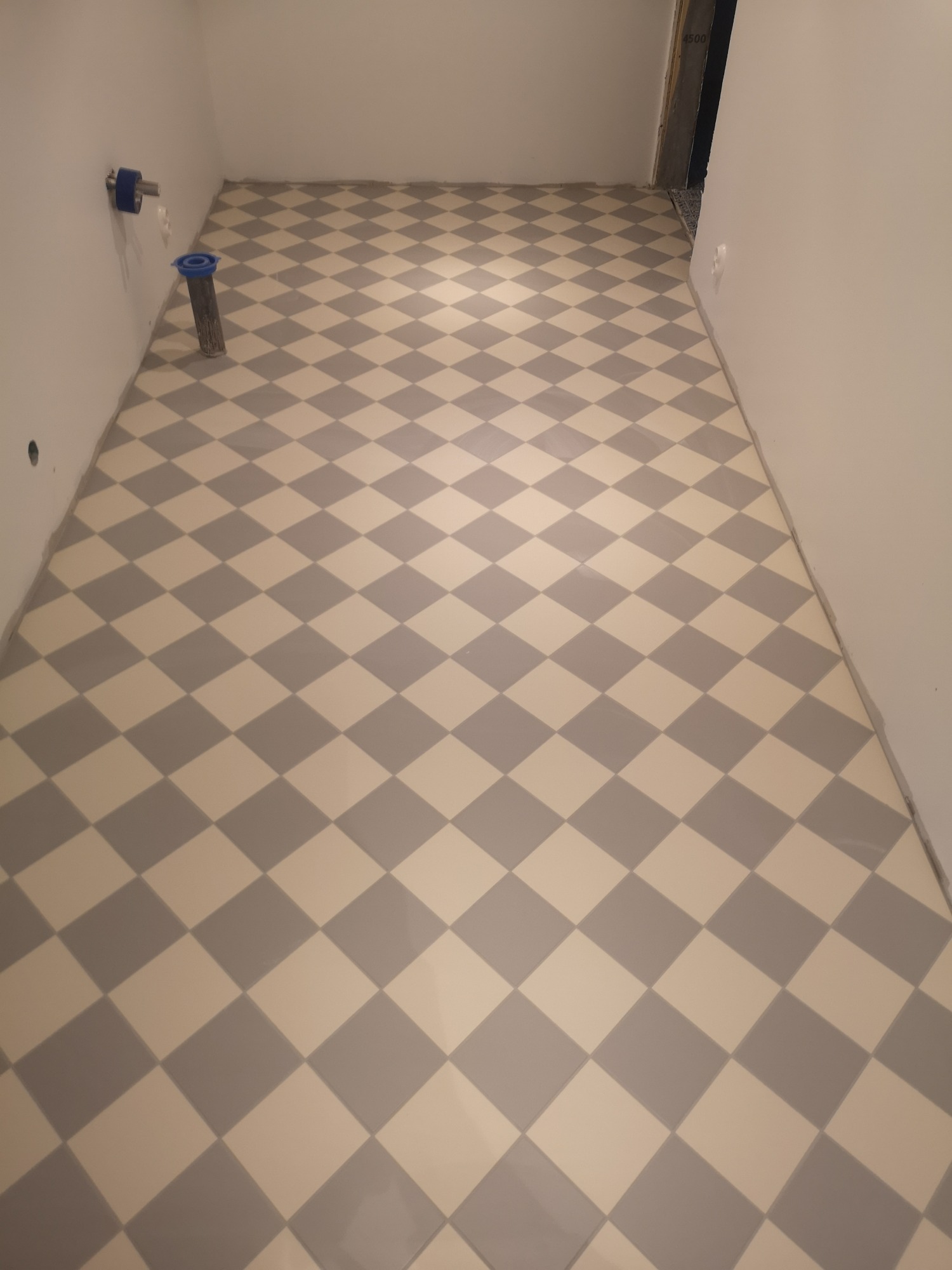 Ukończona podłoga w łazience, wyłożona błyszczącymi, szaro-białymi płytkami w romby, z widocznymi przyłączami hydraulicznymi na ścianie i fragmentem progu drzwiowego.