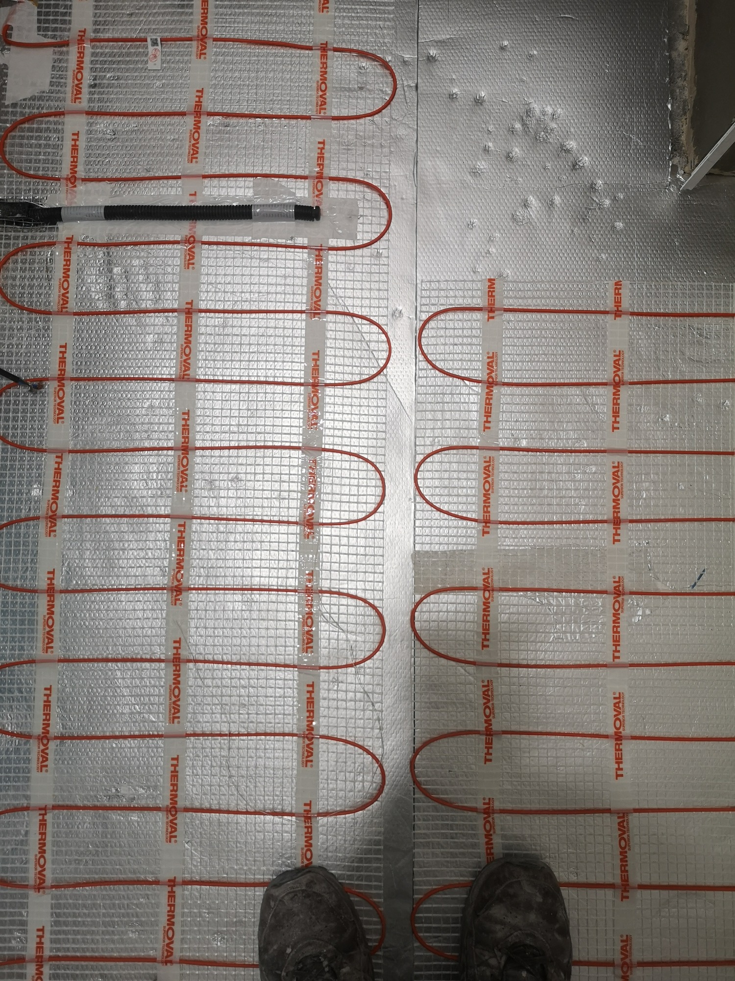 Montaż elektrycznego ogrzewania podłogowego Thermoval na srebrnej folii izolacyjnej, widoczne buty montera w dolnej części kadru.
