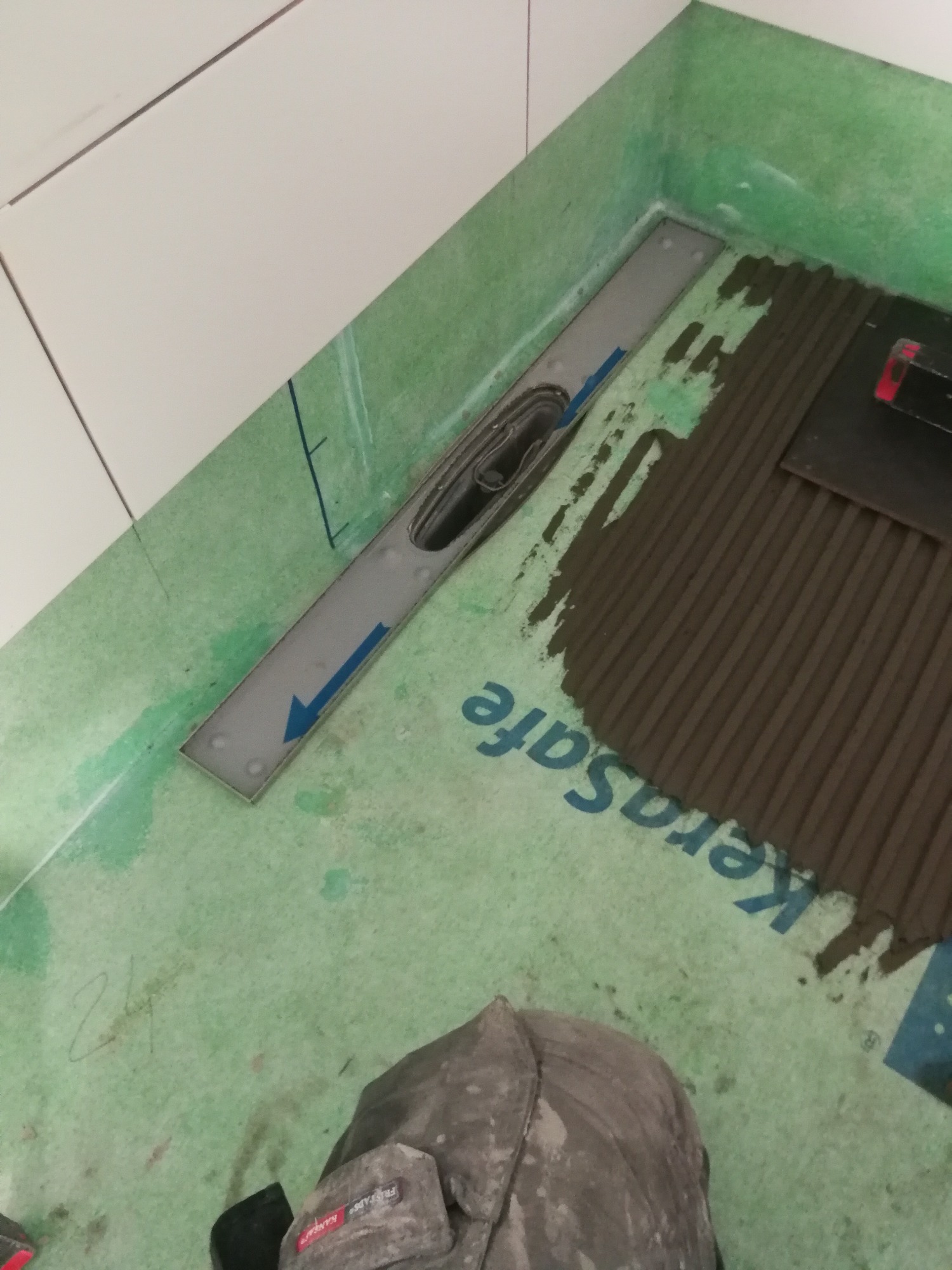 Montaż odpływu liniowego w nowej łazience, widoczna hydroizolacja KeraSafe, fragment ułożonych płytek i narzędzia pracy, perspektywa z góry.