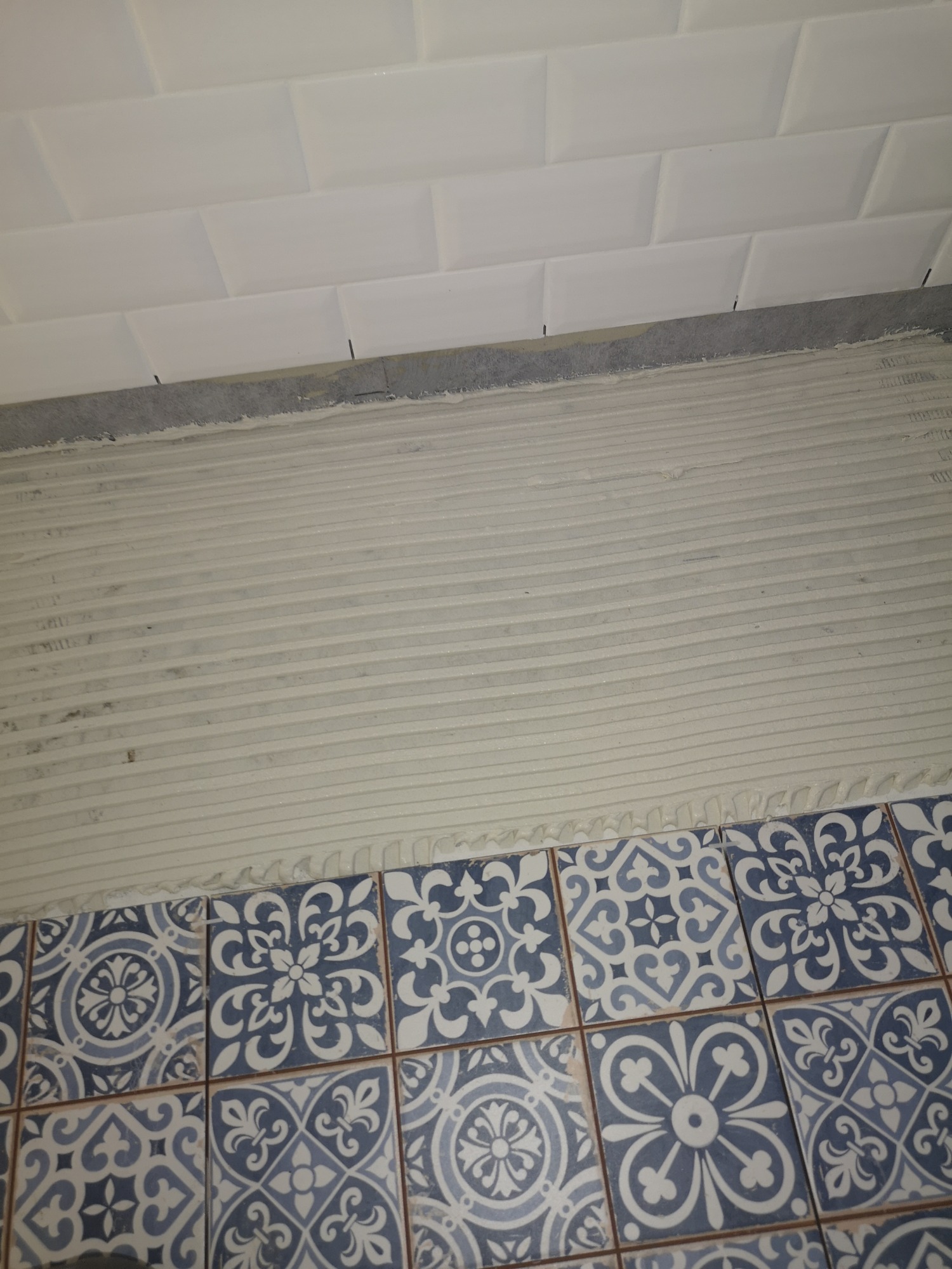 Układanie dekoracyjnych, niebiesko-białych płytek podłogowych na warstwie kleju ząbkowaną pacą, widoczne białe płytki ścienne powyżej.