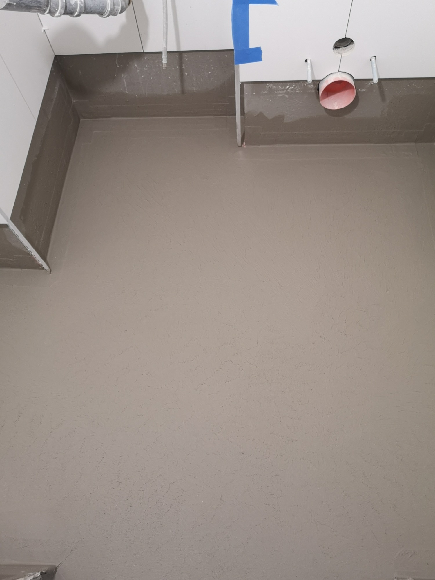 Wyrównana, szpachlowana powierzchnia podłogi w łazience, widoczne rury instalacyjne pod sufitem i fragmenty białych ścian, przygotowane do dalszych prac wykończeniowych.