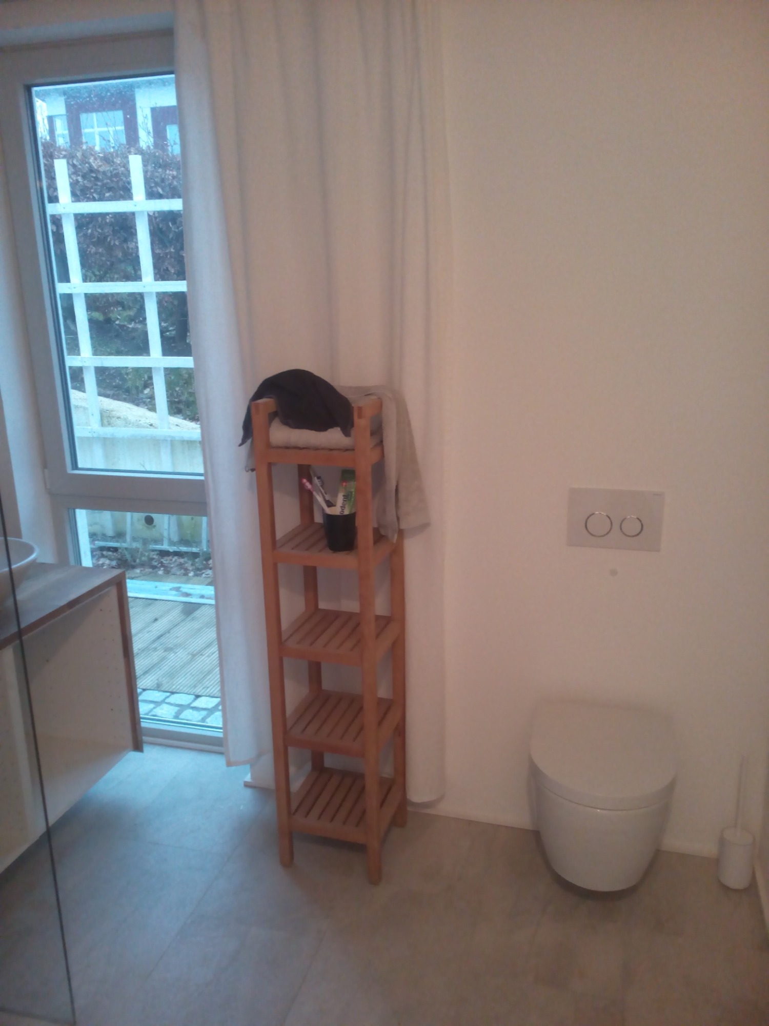 Nowoczesna toaleta z wiszącą muszlą klozetową, drewnianym regałem z akcesoriami i oknem z widokiem na ogród, wykończona w jasnych odcieniach.