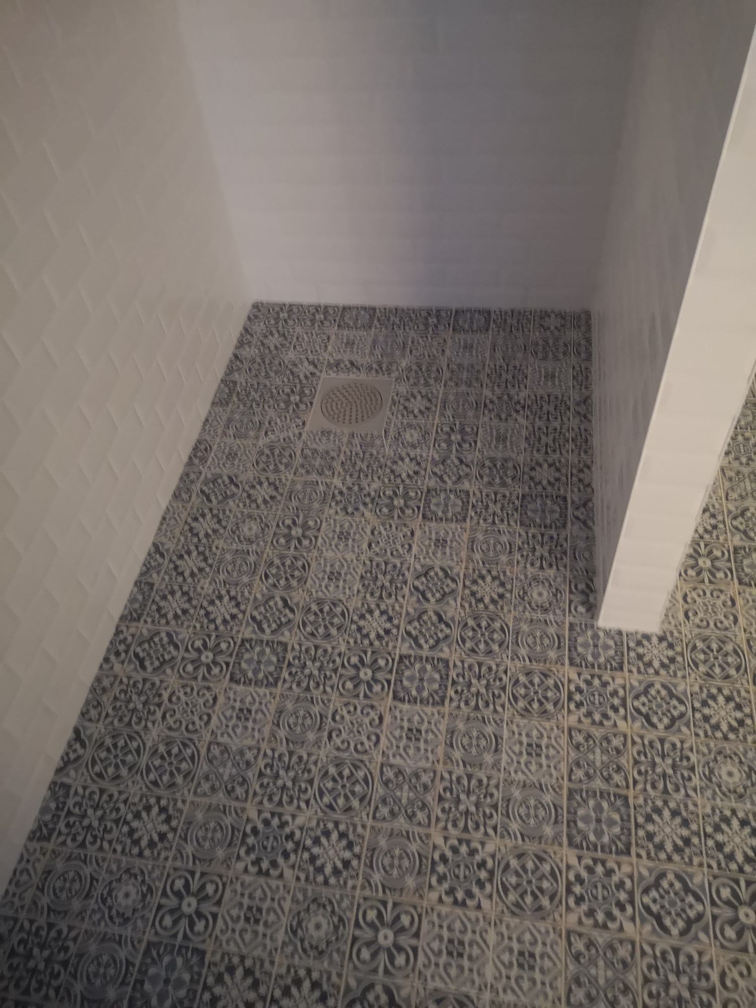 Ułożone, wzorzyste, szaro-niebieskie płytki podłogowe w kabinie prysznicowej z odpływem liniowym, białe ściany wyłożone białą cegiełką.