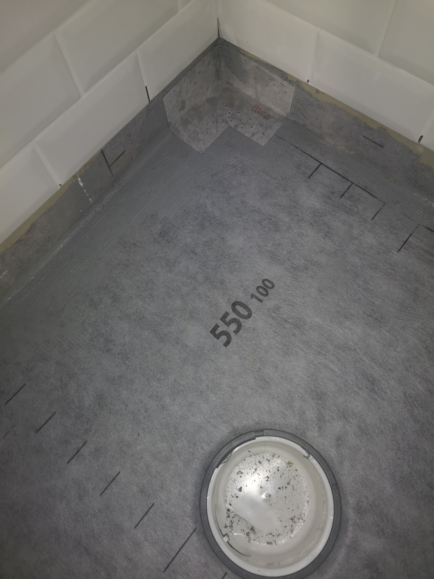 Przygotowanie podłoża pod prysznic: hydroizolacja z widocznym odpływem liniowym, warstwa izolacyjna z nadrukiem '550 100' i zaznaczonymi liniami cięcia, białe płytki na ścianie w tle.