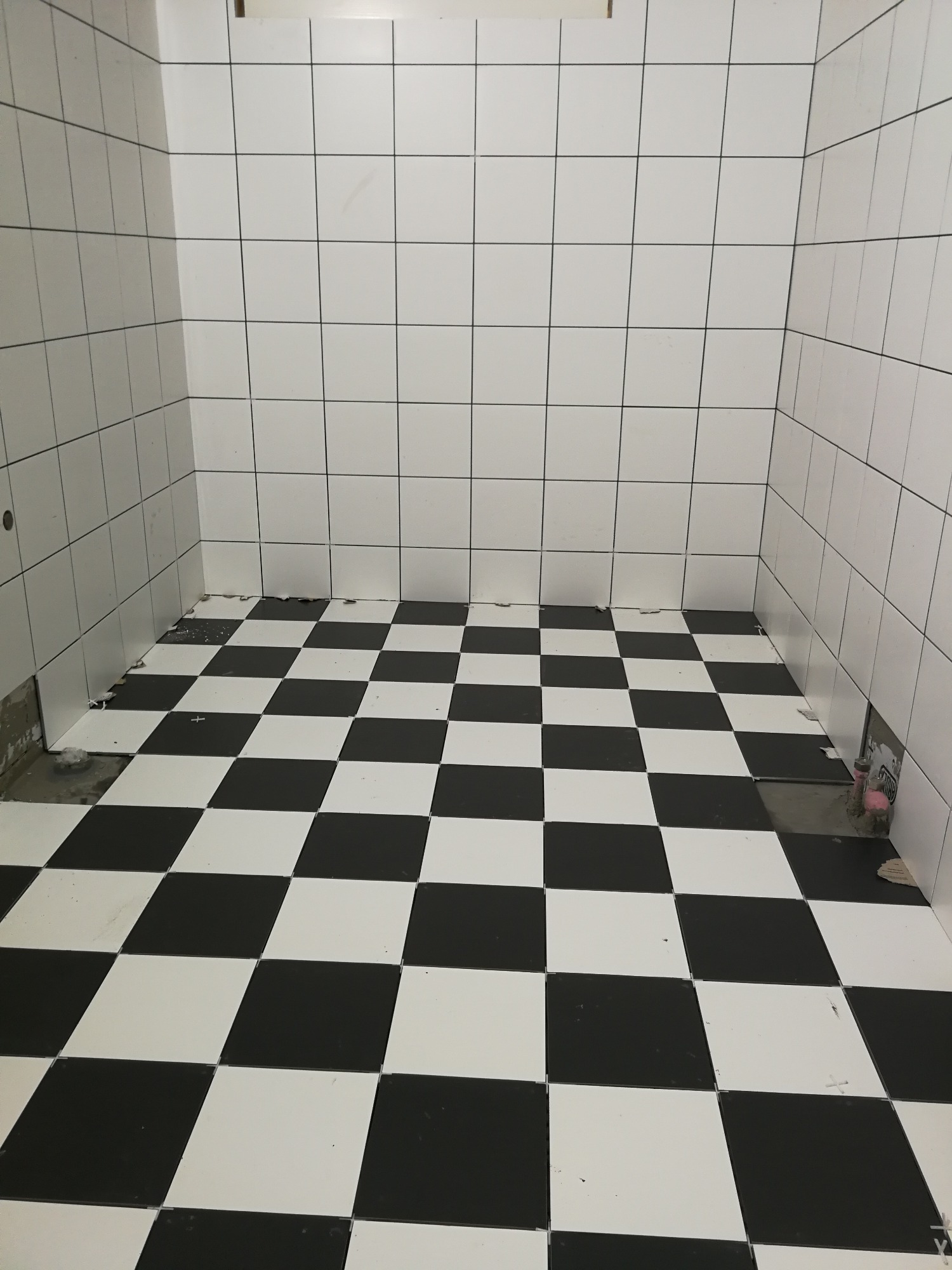 Łazienka w trakcie remontu, białe płytki ścienne i czarno-biała szachownica na podłodze, widoczne ubytki w fugach i brakujące płytki przy ścianie.