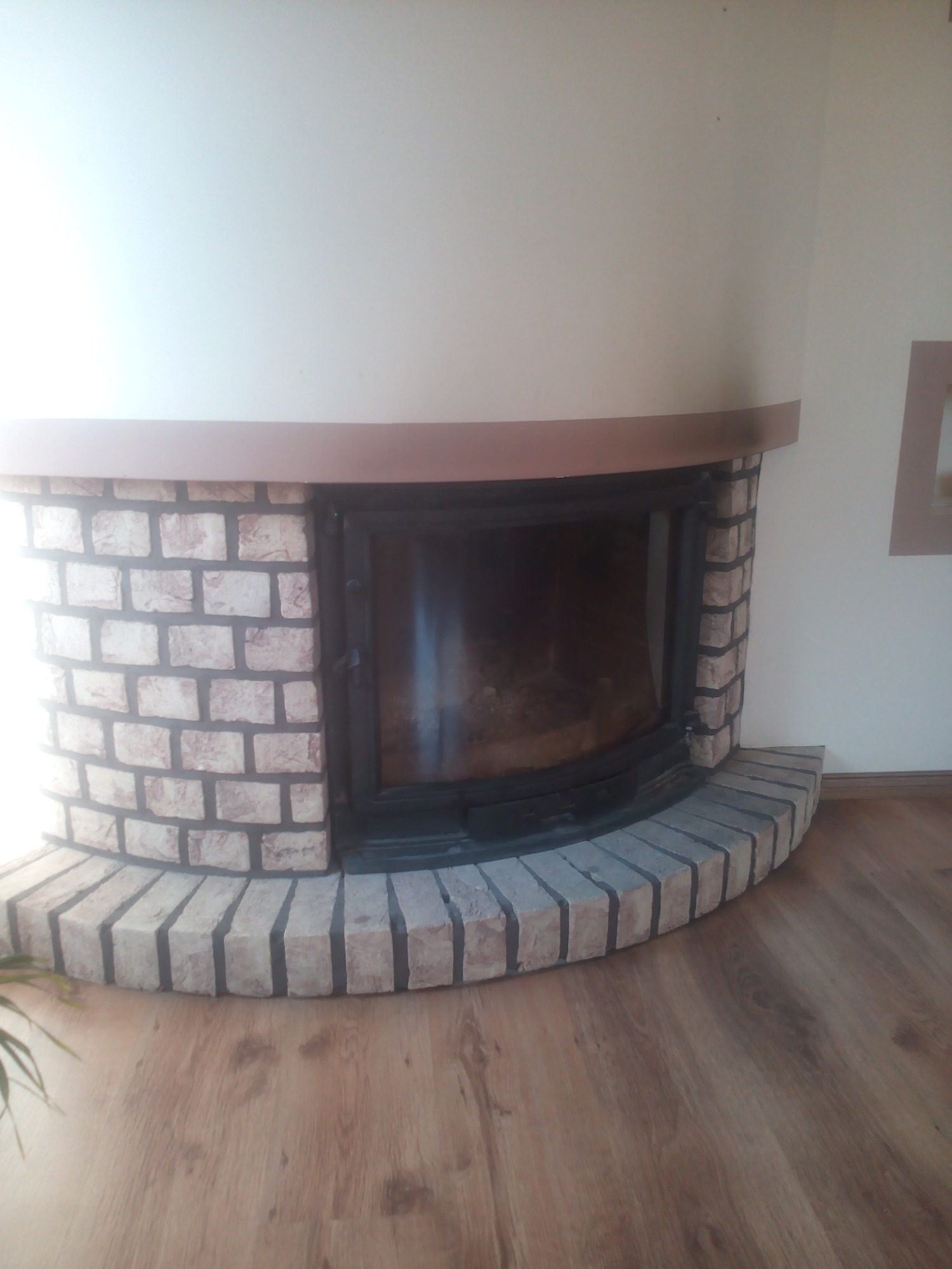 Wykończony kominek z obudową z cegieł w odcieniach beżu i szarości, drewniany blat nad paleniskiem, na podłodze panele imitujące drewno.