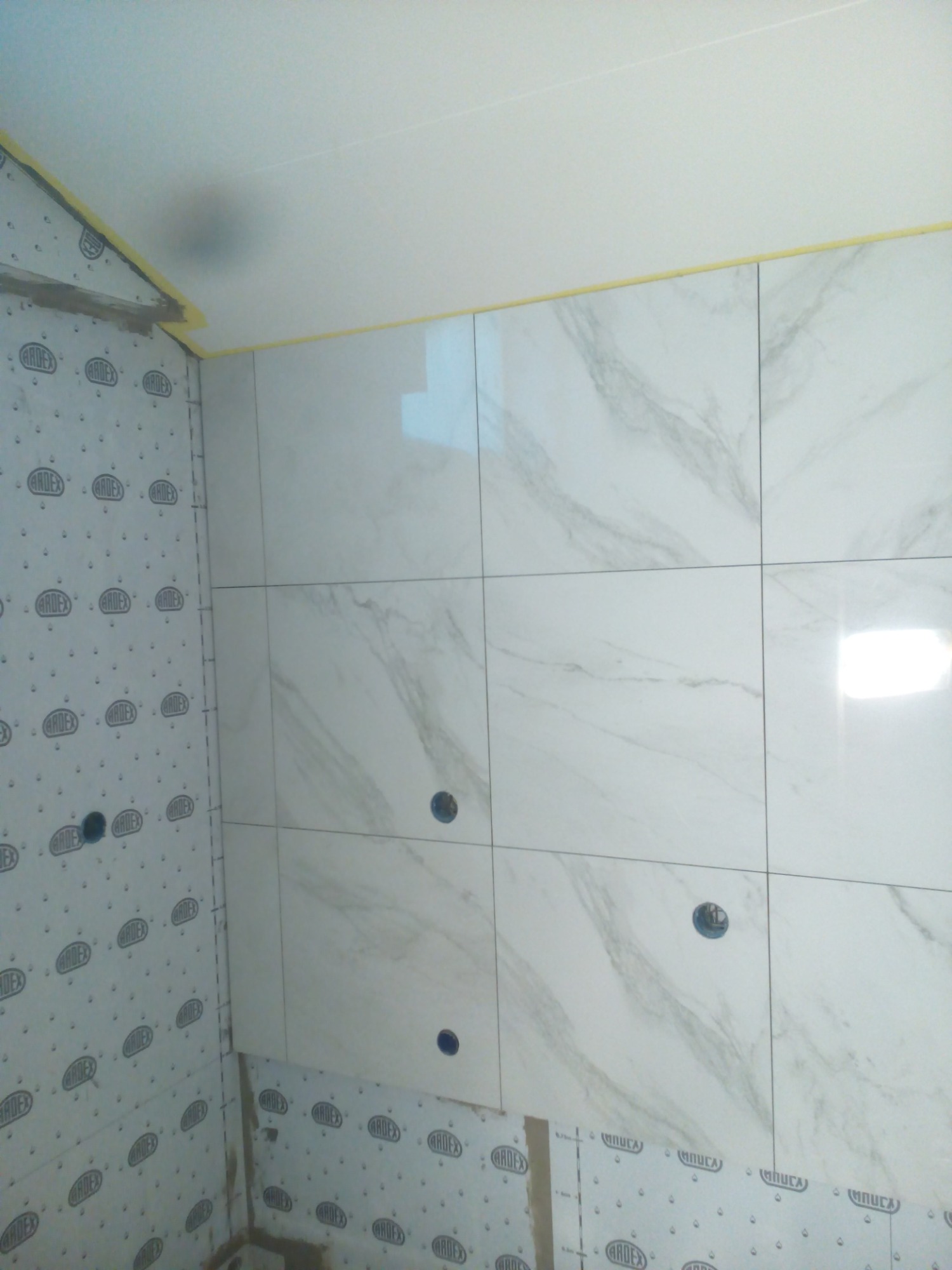 Ściana łazienki w trakcie wykańczania: białe płytki imitujące marmur z wywierconymi otworami na instalacje, przyklejone do płyty montażowej z widocznym logo producenta, zabezpieczone taśmą malarską...