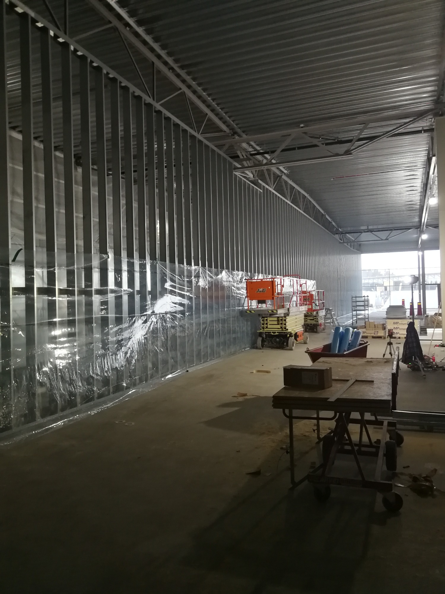 Wnętrze hali w trakcie prac budowlanych: stalowa konstrukcja ścian działowych, zabezpieczona folią, podnośniki nożycowe JLG w tle, stół roboczy na pierwszym planie.