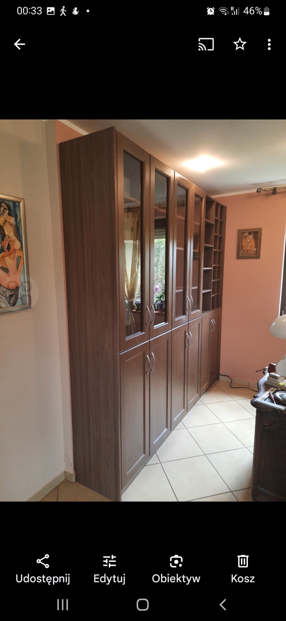 Brązowa szafa wnękowa z przeszklonymi drzwiami i otwartymi półkami, dopasowana do wnętrza. Widoczne detale wykończenia i oświetlenie w pomieszczeniu.