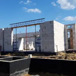 Wznoszenie ścian parteru z betonu kom&oacute;rkowego na zaizolowanym fundamencie, widoczna zbrojenie nadproży i podpory pionowe, w tle błękitne niebo z chmurami.