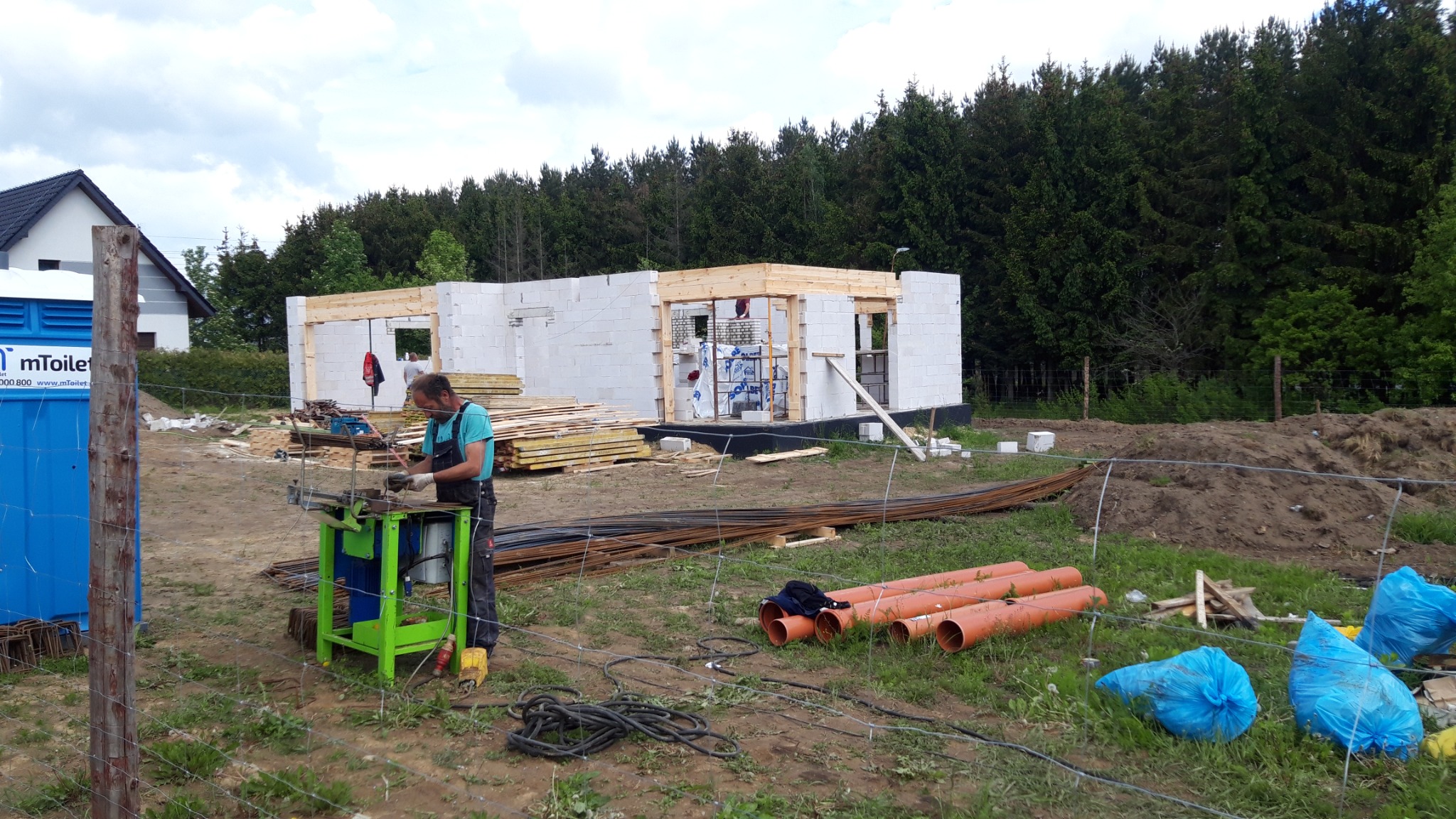 Budowa domu jednorodzinnego z białych bloczków: pracownik na pierwszym planie obrabia stal zbrojeniową przy użyciu zielonej maszyny. W tle widoczna konstrukcja domu z drewnianymi nadprożami, stosy...