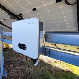 Biały falownik SOFAR zamontowany pod panelami słonecznymi na metalowej konstrukcji, widoczne okablowanie i elementy montażowe, w tle trawa i częściowo widoczny budynek.