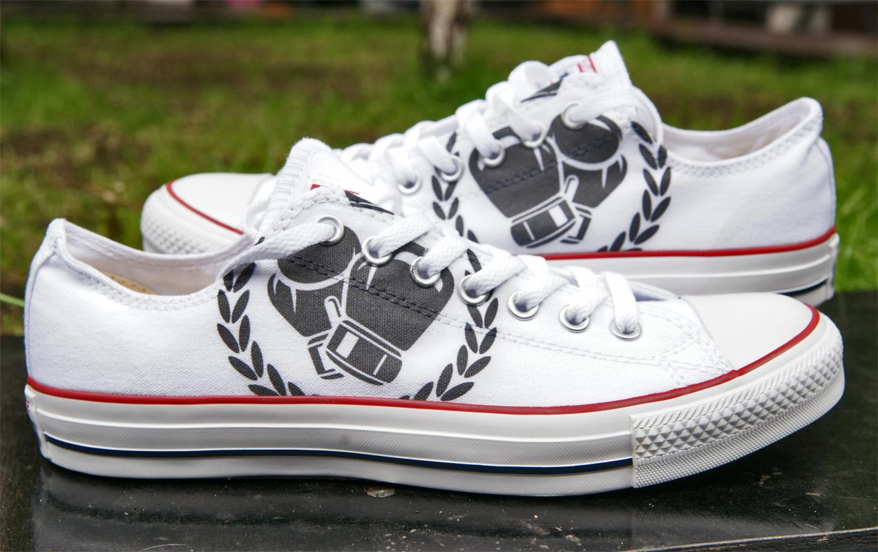 Białe trampki z nadrukiem bokserskich rękawic otoczonych wieńcem laurowym, leżące na ciemnej powierzchni, z zielonym tłem. Unikatowy wzór na obuwiu.