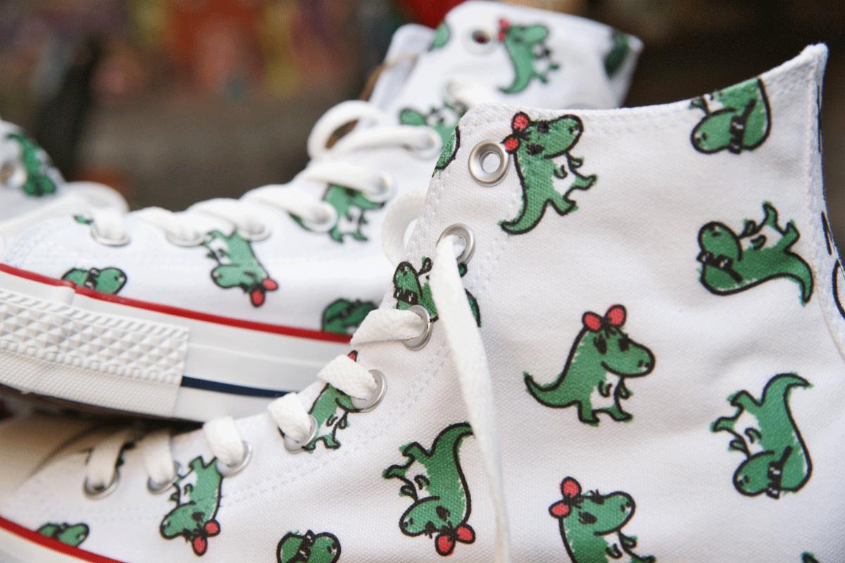 Białe trampki z nadrukiem zielonych dinozaurów z kokardkami, widoczne sznurówki i podeszwa z czerwonym paskiem, unikalny wzór na obuwiu.