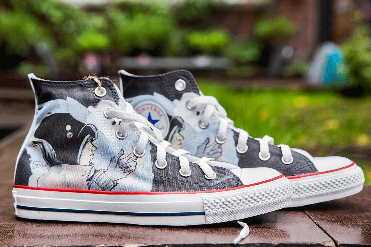 Dwa trampki Converse z unikalnym nadrukiem przedstawiającym astronautów w stylu vintage, ustawione na drewnianej powierzchni na tle zieleni.