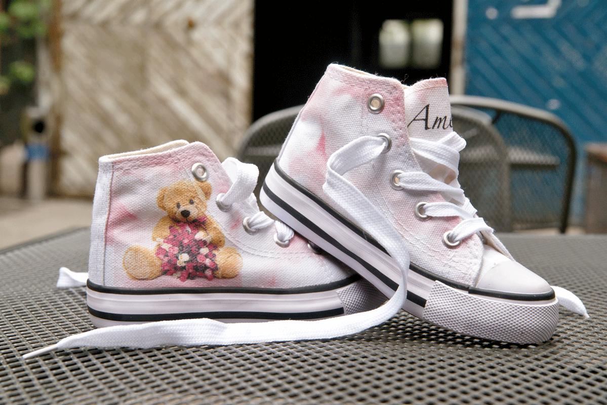 Białe trampki dziecięce z nadrukiem misia trzymającego kwiaty, leżące na metalowym stoliku, z widocznymi sznurówkami i detalami wykończenia.