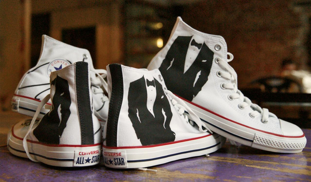 Białe trampki Converse z czarnym nadrukiem smokingu, ułożone na fioletowym stole. Widoczne detale sznurowadeł i podeszwy.