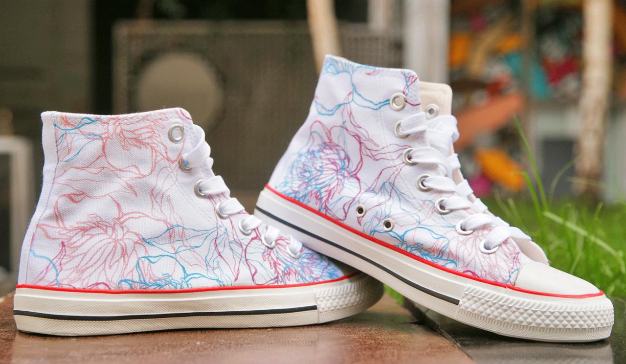 Białe, sznurowane trampki z wysoką cholewką, zdobione wzorem kwiatowym w odcieniach różu i błękitu, leżące na drewnianej powierzchni.