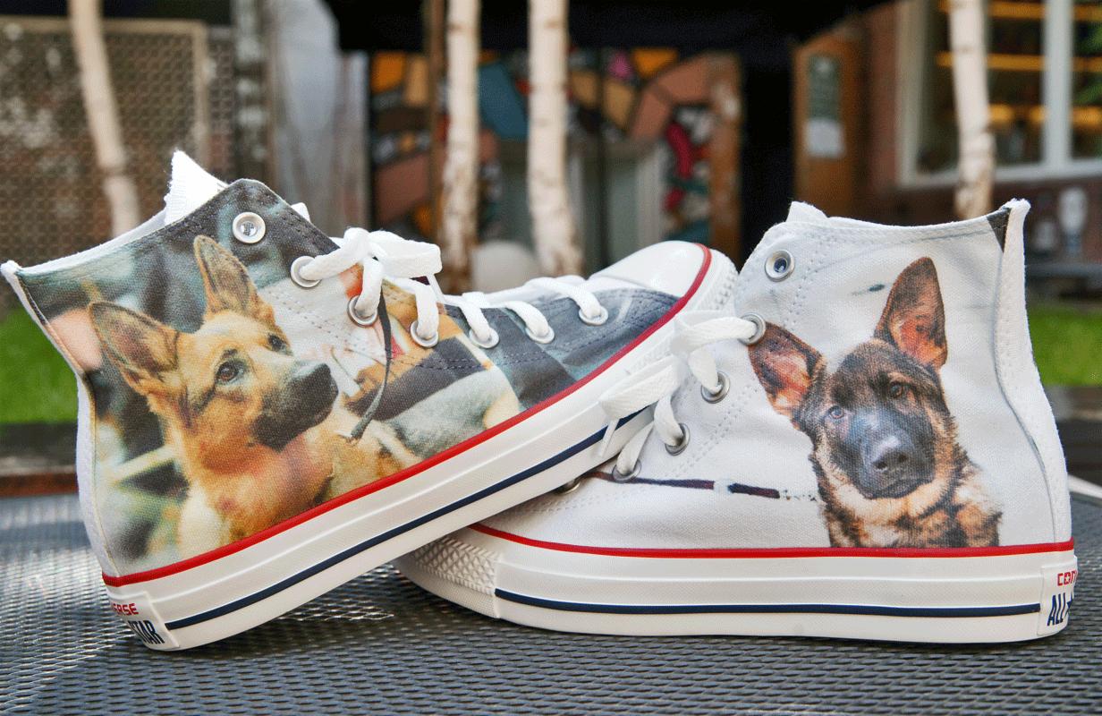 Białe trampki z nadrukiem portretów owczarka niemieckiego, ustawione na metalowej powierzchni, w tle rozmyte drzewa i budynek.