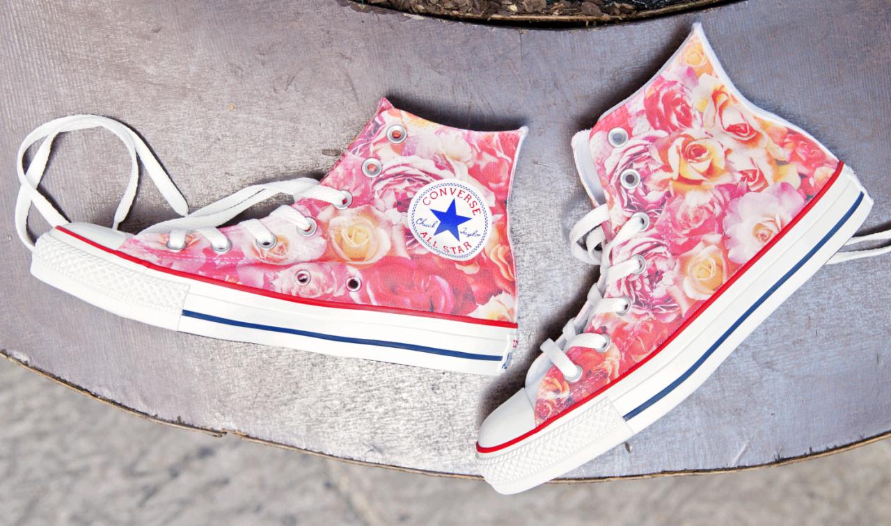 Dwa wysokie trampki Converse z nadrukiem róż, leżące na szarej, metalowej powierzchni. Jeden but z boku, drugi z przodu, sznurówki białe.