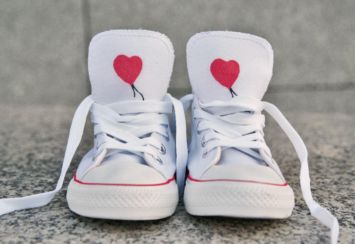Białe trampki z czerwonym sercem zamiast logotypu, z białymi sznurówkami, na szarym tle. Widok z przodu na parę butów. Czerwona lamówka na podeszwie.