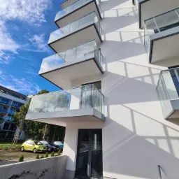 Nowoczesny budynek mieszkalny z balkonami z przezroczystymi panelami, widoczne zaparkowane samochody i błękitne niebo z chmurami.