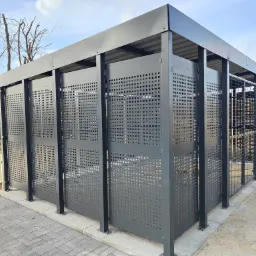 Nowoczesna, metalowa wiata rowerowa z perforowanymi ścianami, umieszczona na nowo wybrukowanym chodniku, z widocznym fragmentem terenu budowy w tle.