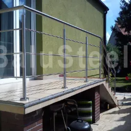 Zewnętrzna platforma z wyjściem balkonowym i schodami, zabezpieczona stalową konstrukcją z poziomymi poprzeczkami, widok na ogród z kwitnącymi kwiatami.