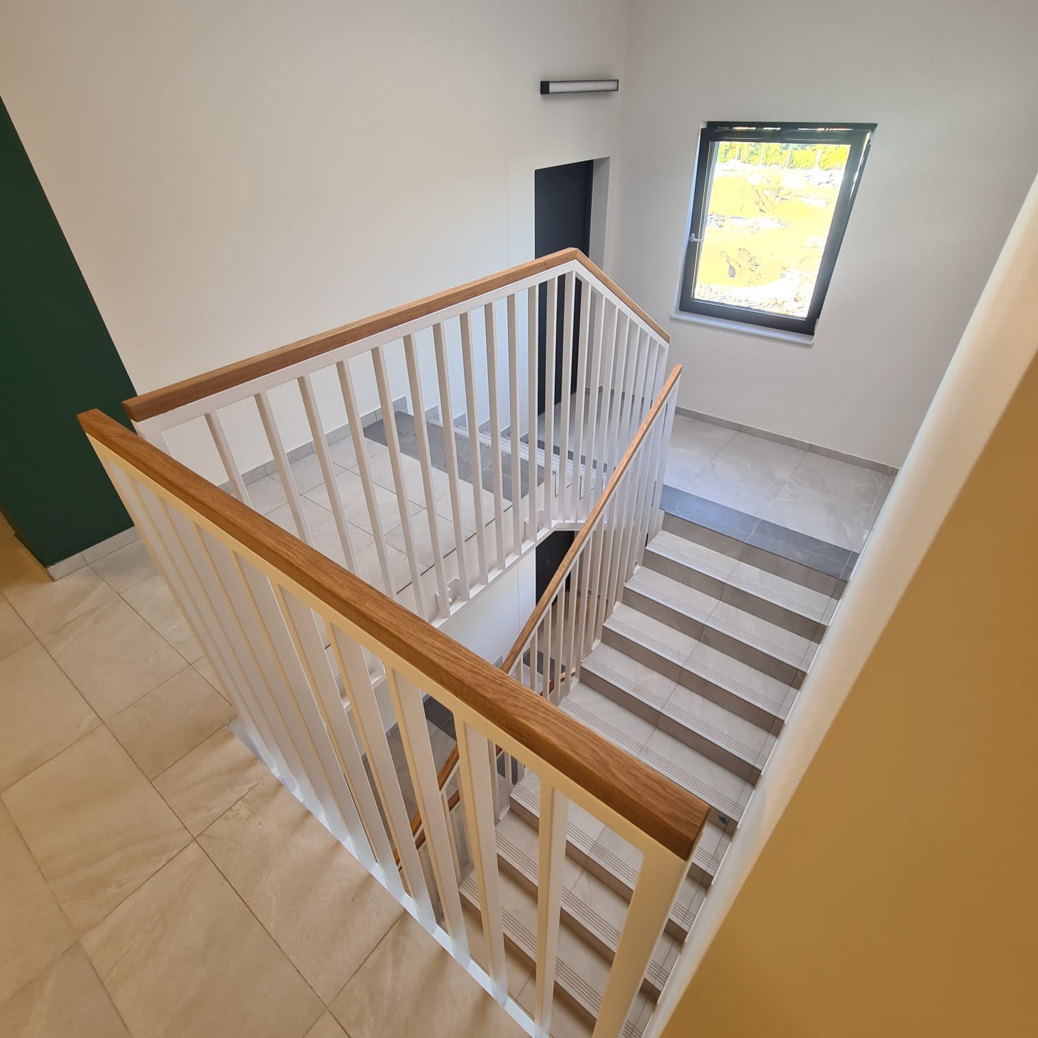 Wysoki kąt widzenia na schody wewnętrzne z białymi pionowymi barierkami i drewnianym poręczą, widok z góry na dół, neutralna kolorystyka ścian i podłogi, okno w tle.