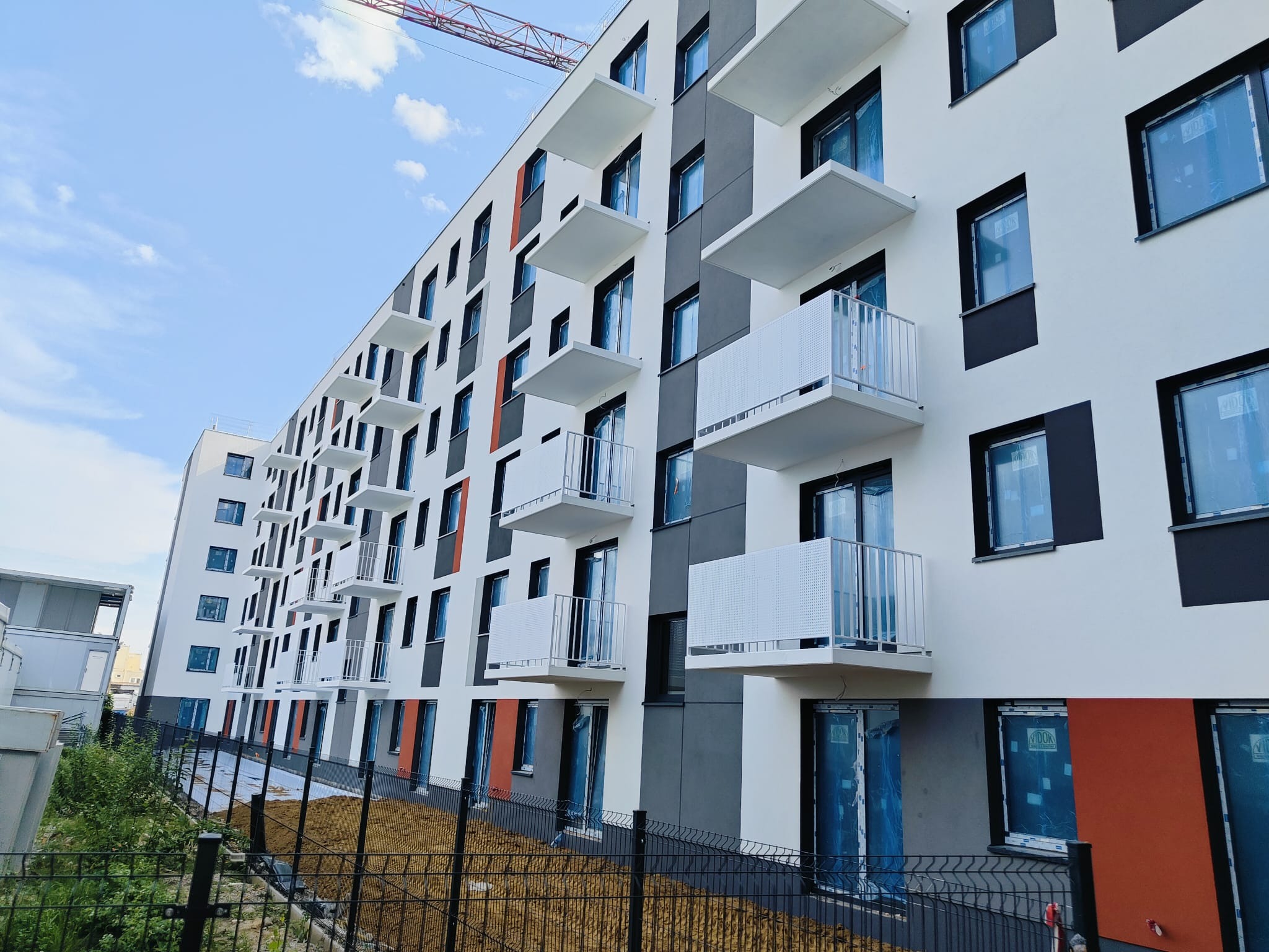 Nowoczesny budynek mieszkalny z balkonami zabezpieczonymi ażurowymi barierkami, widok z dołu na elewację w kolorach bieli, szarości i oranżu, plac budowy w tle.