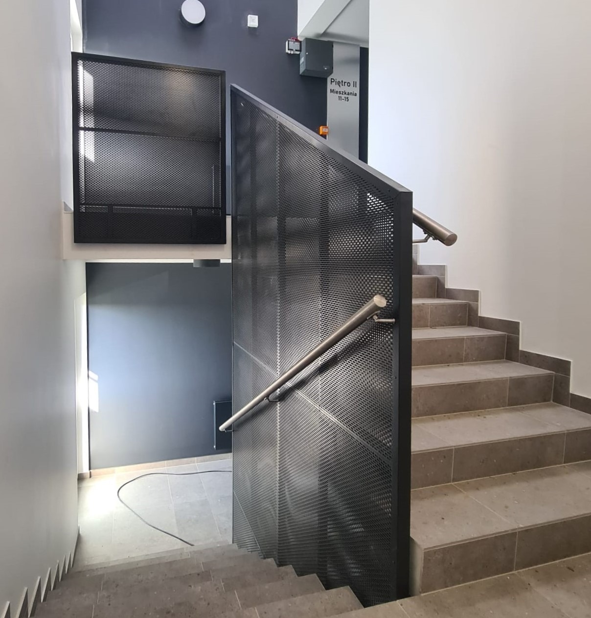 Klatka schodowa z metalową ażurową balustradą i poręczą ze stali nierdzewnej, widok z góry na schody wyłożone płytkami.