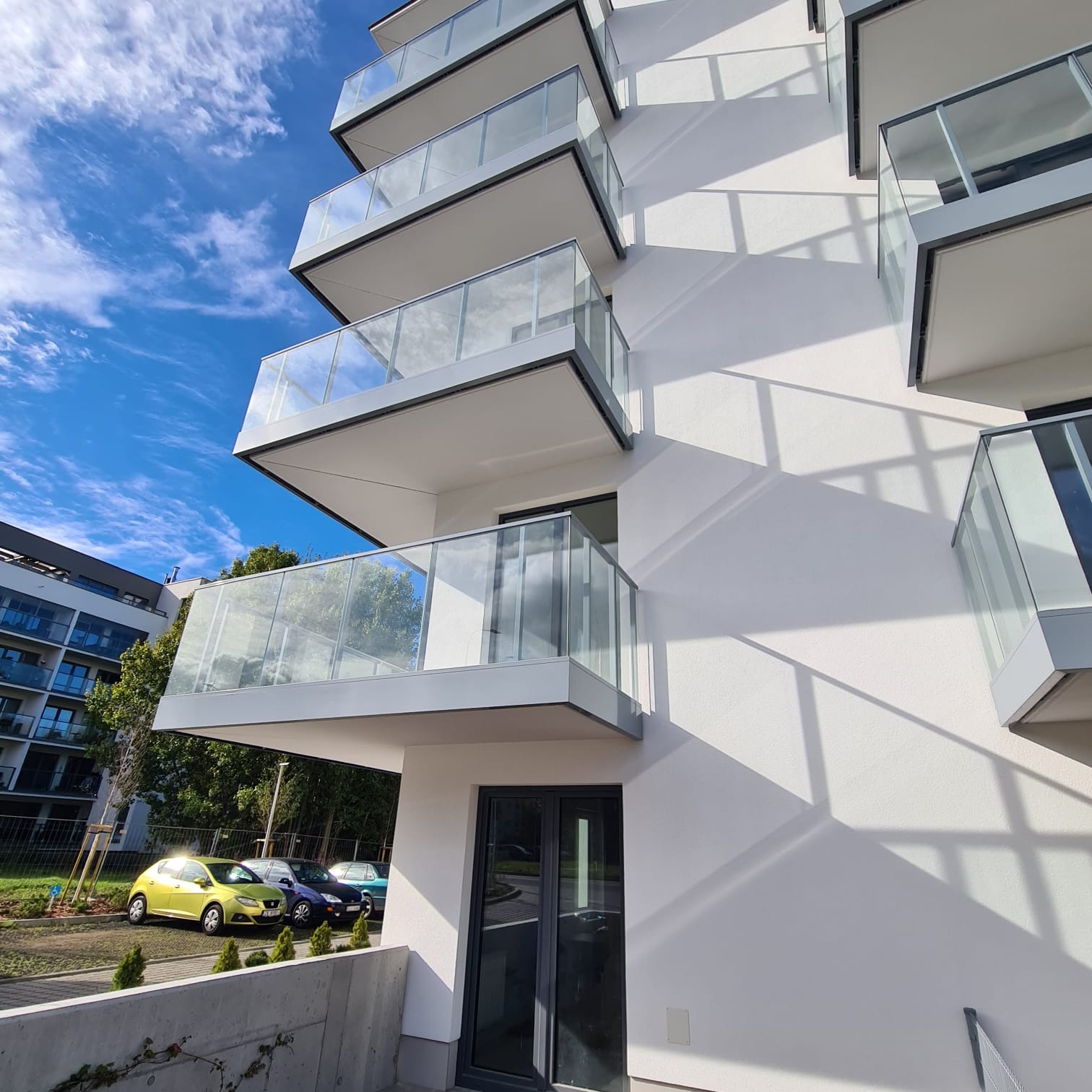 Nowoczesny budynek mieszkalny z balkonami z przezroczystymi panelami, widoczne zaparkowane samochody i błękitne niebo z chmurami.